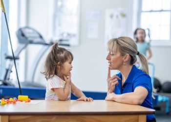 A female therapist sits with a sweet little girl as a table as they work on her speech together.  The Therapist is dressed professionally and is pointing to her mouth to help show the little girl how to form her words.